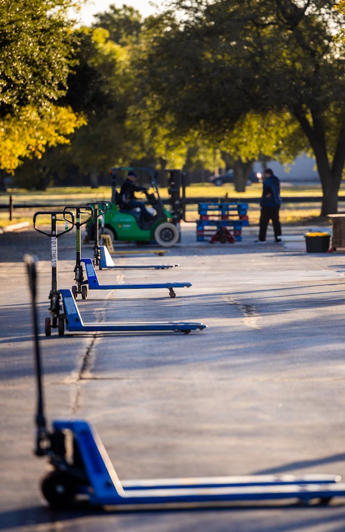 Pallet jacks and forklift in an outdoor industrial setting with trees and sunlight.
